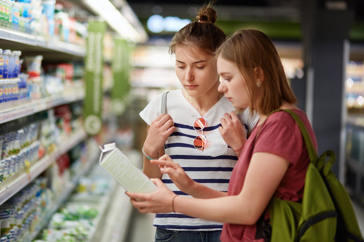 Two pretty female sisters go shopping together, stands in grocer`s shop, select fresh milk in paper container, read label, carry rucksacks, have serious expressions. People and commerce concept; Shutterstock ID 1119506393
